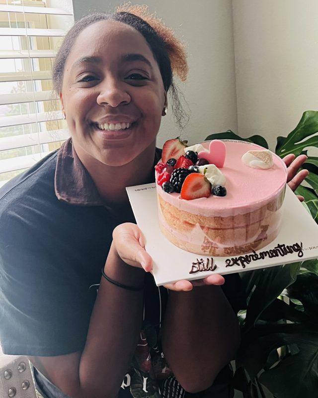 Jai holding a pink cake with "still experimenting!" written on the plate