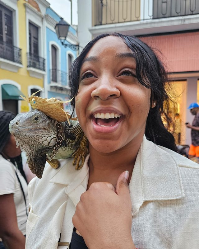 Jai laughing in delight with an iguana in a tiny straw hat perched on her shoulder
